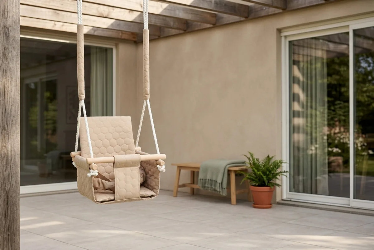 Beige babygynge hængt op i en pergola på en flisebelagt terrasse med salviegrøn plaid og terrakottakrukke
